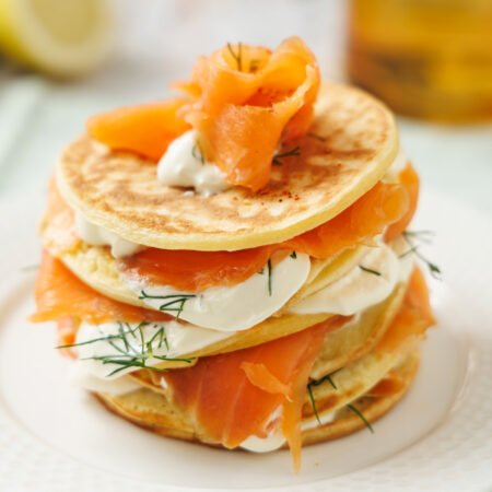 Millefeuille de blinis avec des tranches de truite fumée et crème fouettée à l'aneth et au citron, dressé dans une assiette blanche